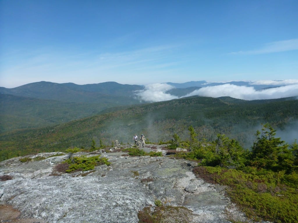 Two hikers climb a steep rocky slope with a mountain range in the background.