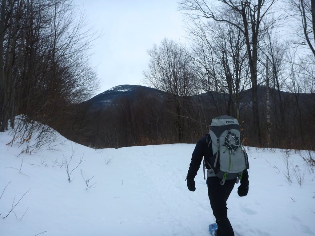 A lone hiker walks down a snowy trail towards a distant mountain range.