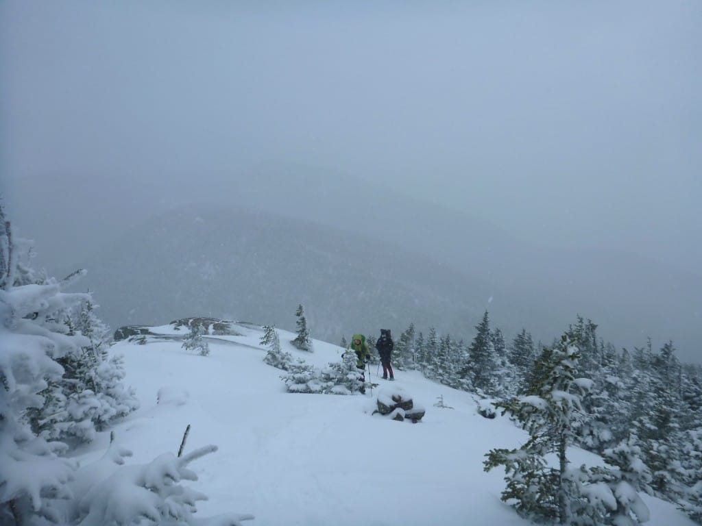 Two hikers stand on a snowy ridge, surrounded by fog.