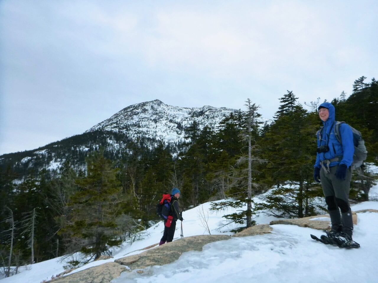 Two hikers stand in front of a view of a snowy mountain and evergreen forest.