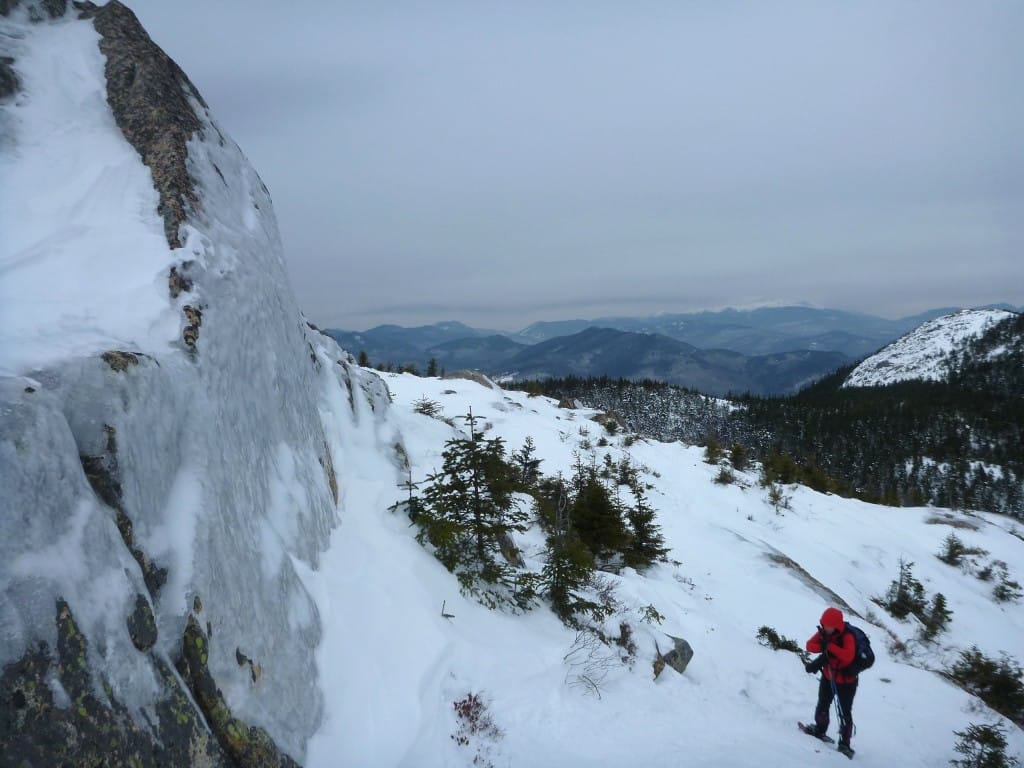A hiker on snowy ground stands next to an imposing cliff.