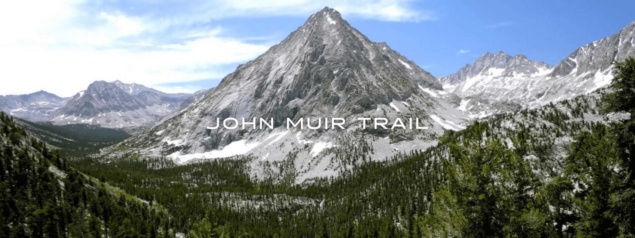 The John Muir Trail offers a commanding view of a rocky mountain peak with patchy snow surrounded by an evergreen forest.
