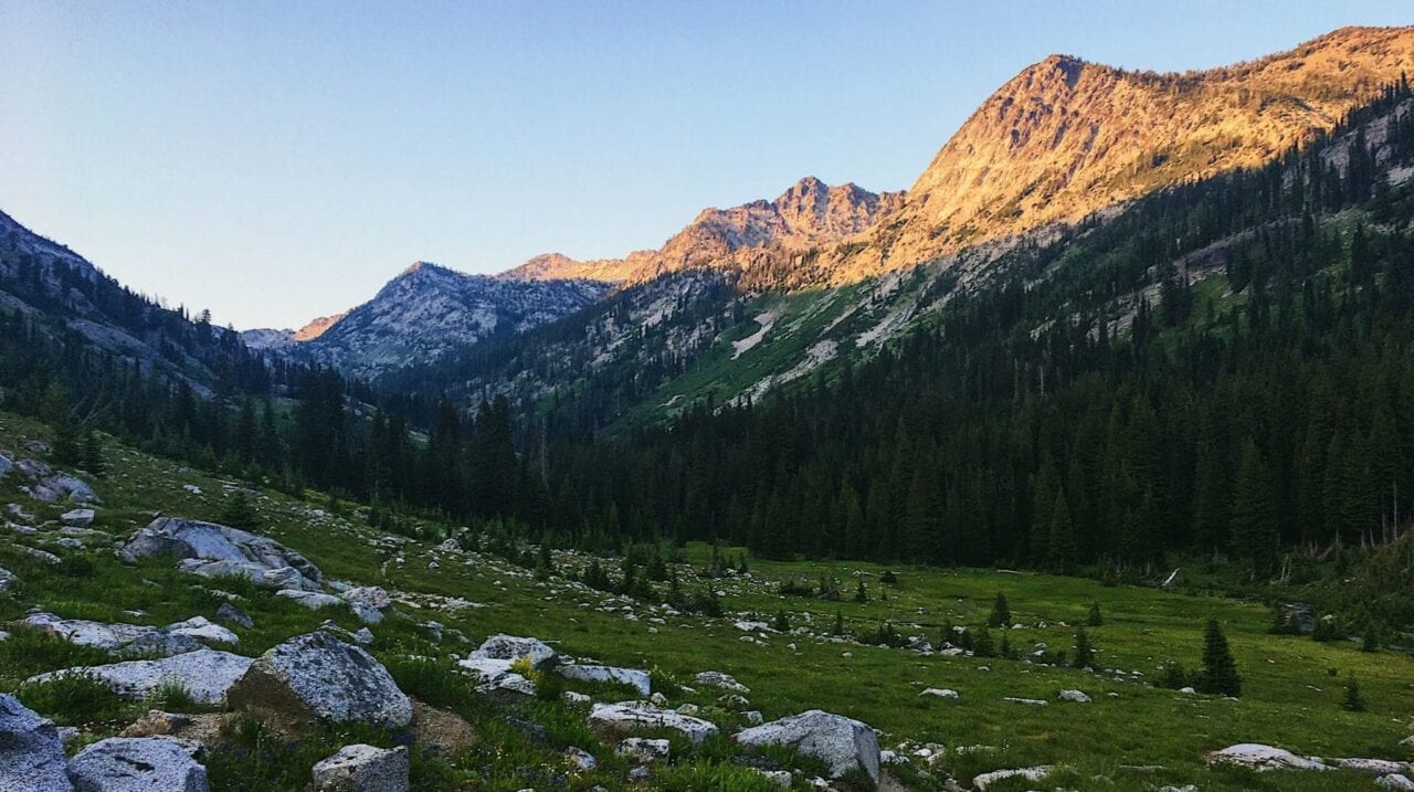 A sunset trows shadows over a rocky green valley surrounded by tall mountain peaks.