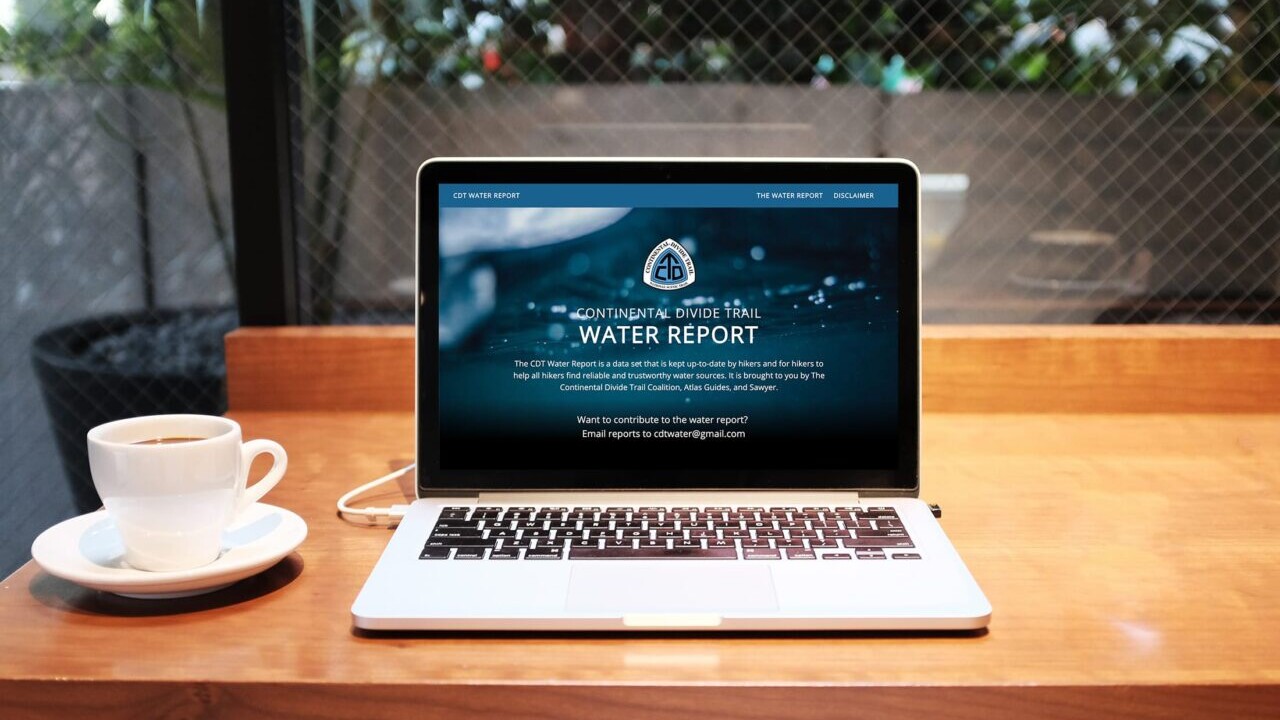 A laptop sits on the table next to a cup of coffee and its screen shows the Continental Divide Trail Water Report website.