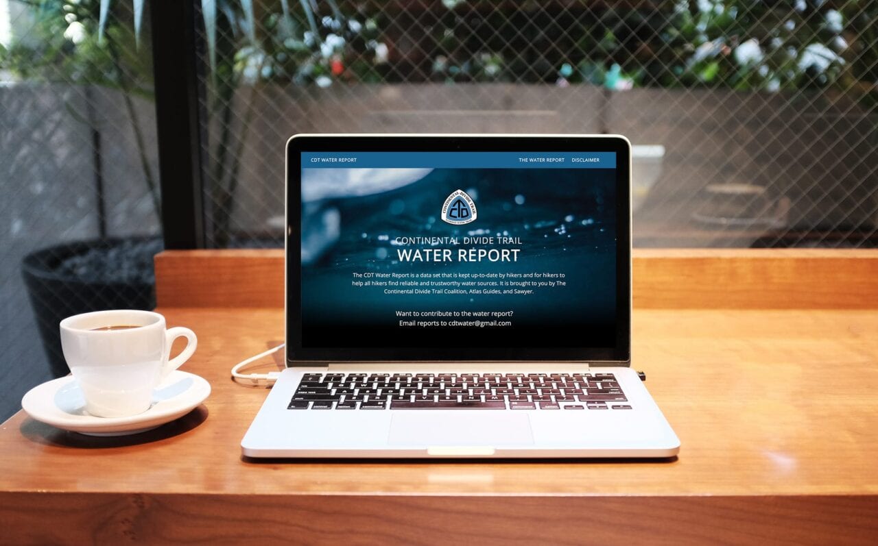 A laptop sits on the table next to a cup of coffee and its screen shows the Continental Divide Trail Water Report website.
