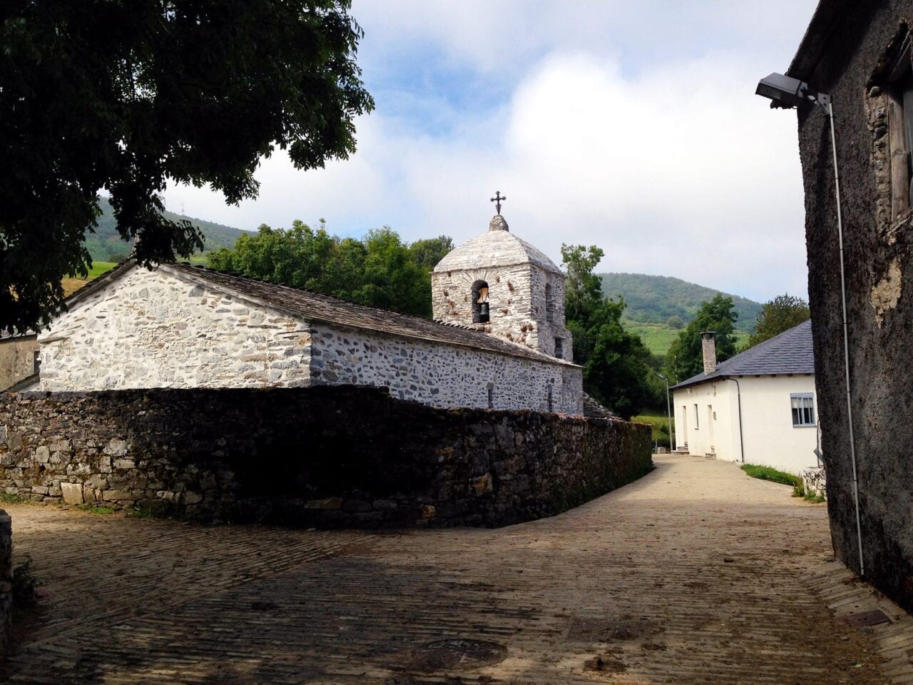 The Camino de Santiago winds through a small town on cobblestone streets with an old, white stone church on the left and green rolling hills in the background.