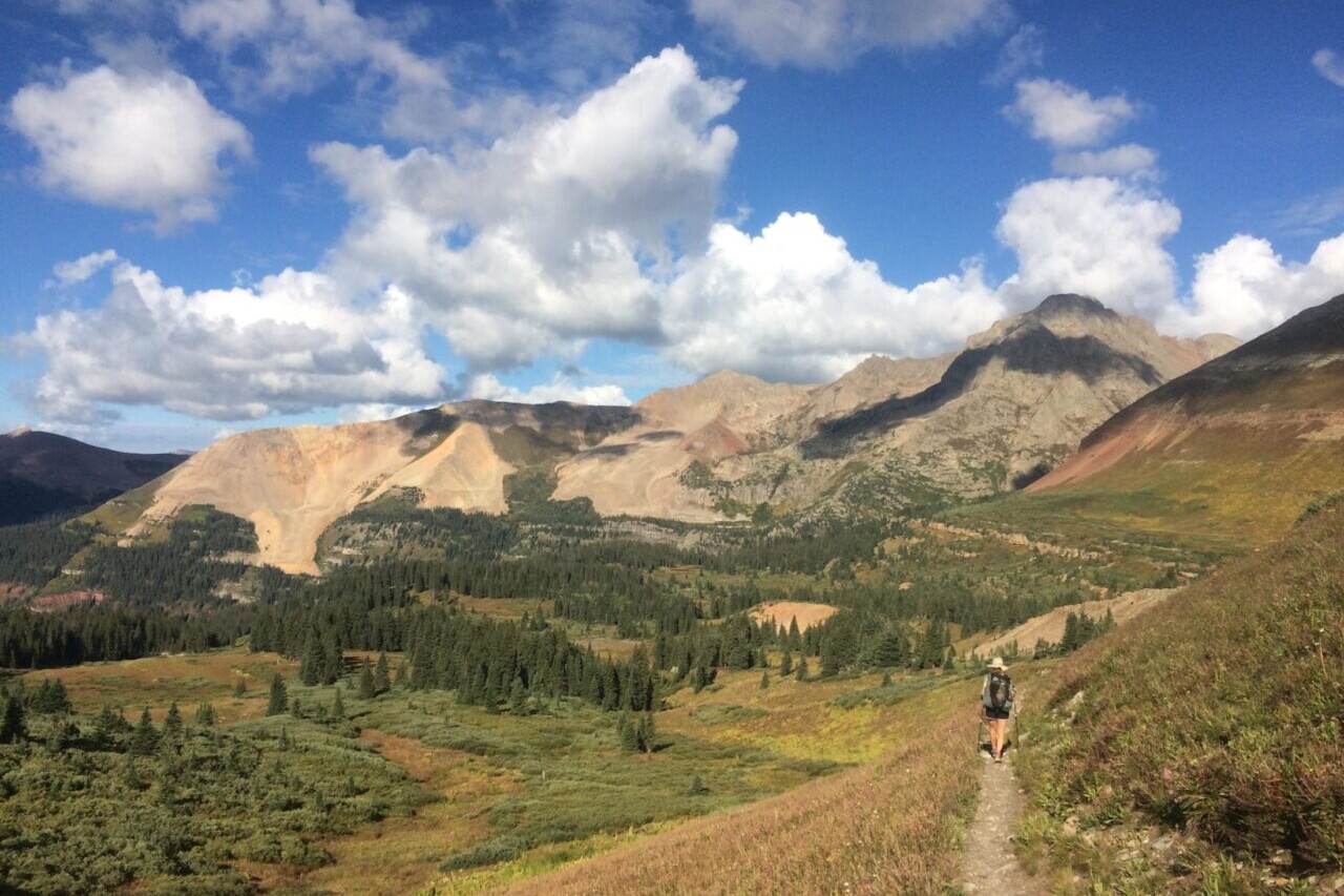 A trail winds around a golden hillside with rocky mountains in the distance.