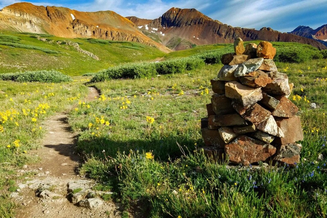 A trail winds through a green meadow past a rock cairn with rocky mountains in the distance.