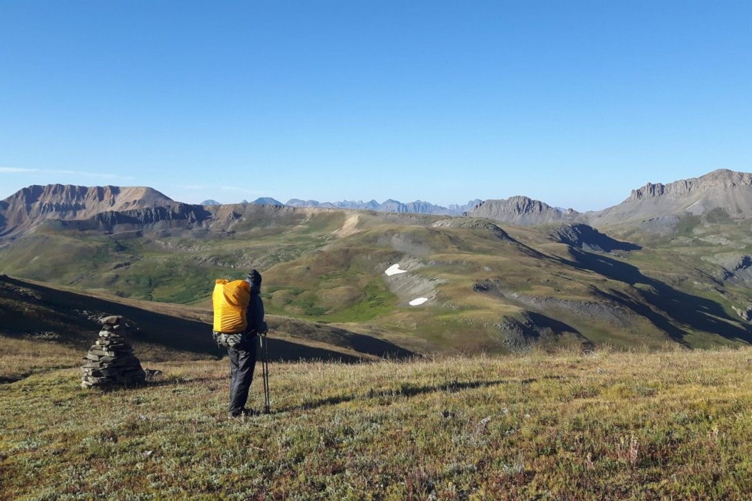 A hiker stands on a hill next to a cairn looking at distant rocky mountains.