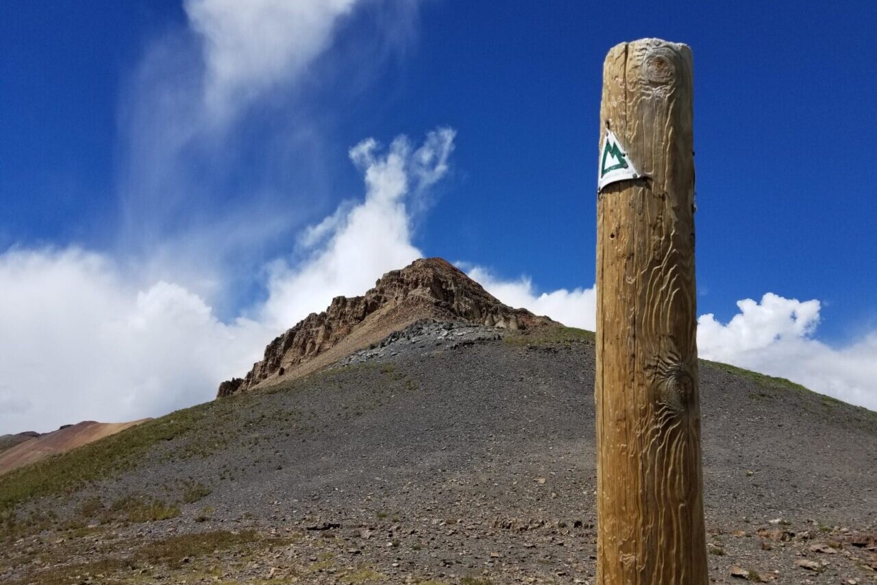 A trail sign stands in front of a rocky mountain peak.