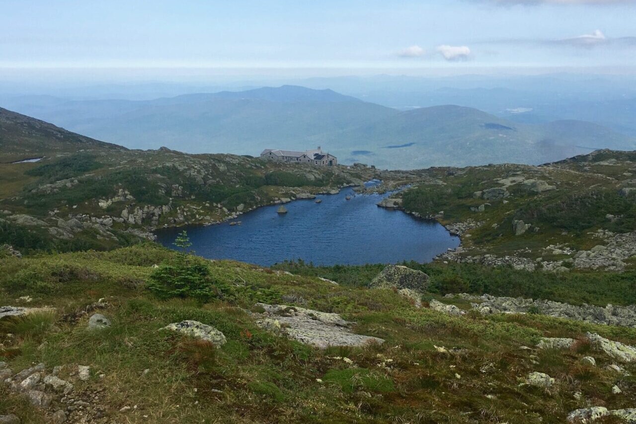 A pond on a rocky ridge in a green field has mountains in the distance.