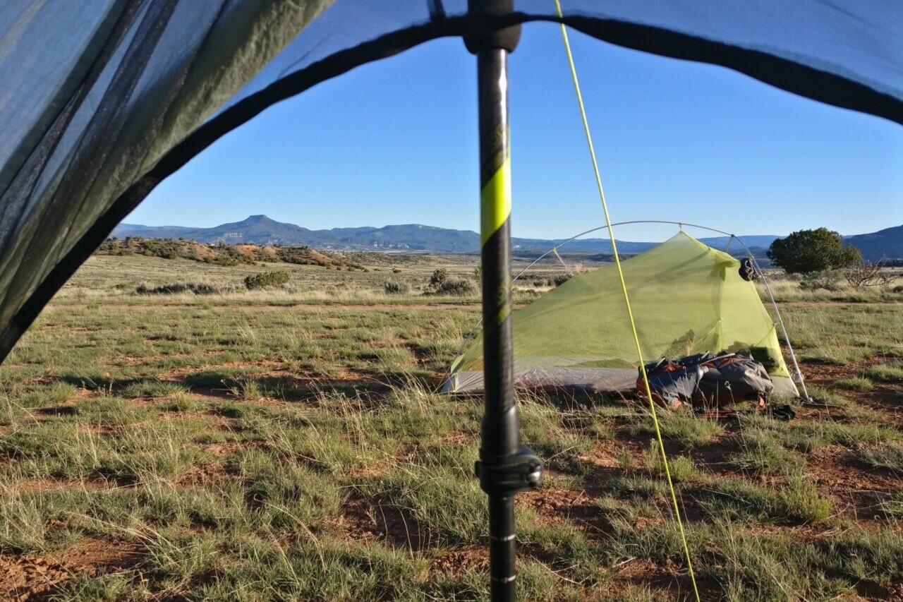 The view from inside a tent shows another tent in an open field and distant mountains.