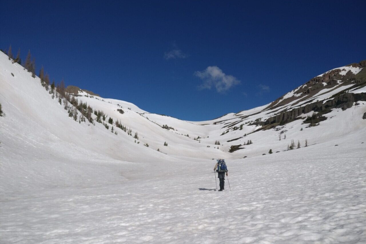A hiker crosses a snowfield towards a pass.