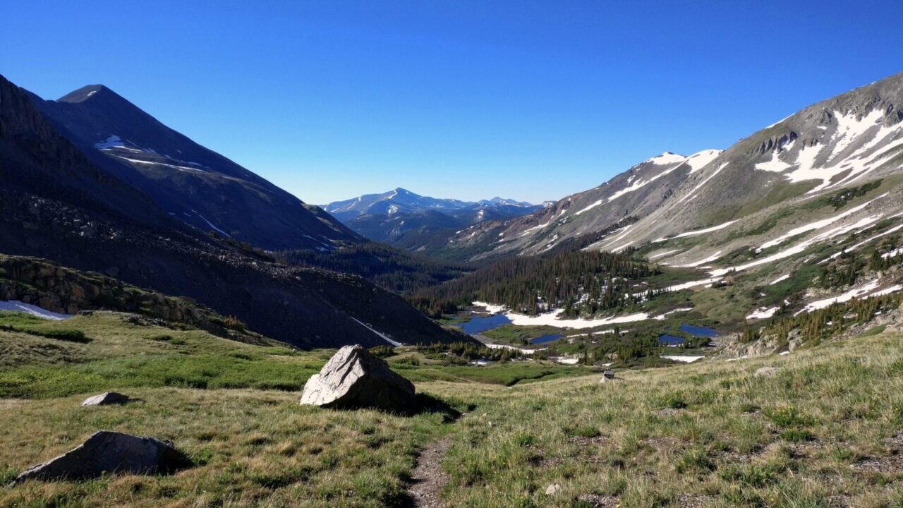 A trail leads through a grassy meadow to distant blue lakes and tall mountains.