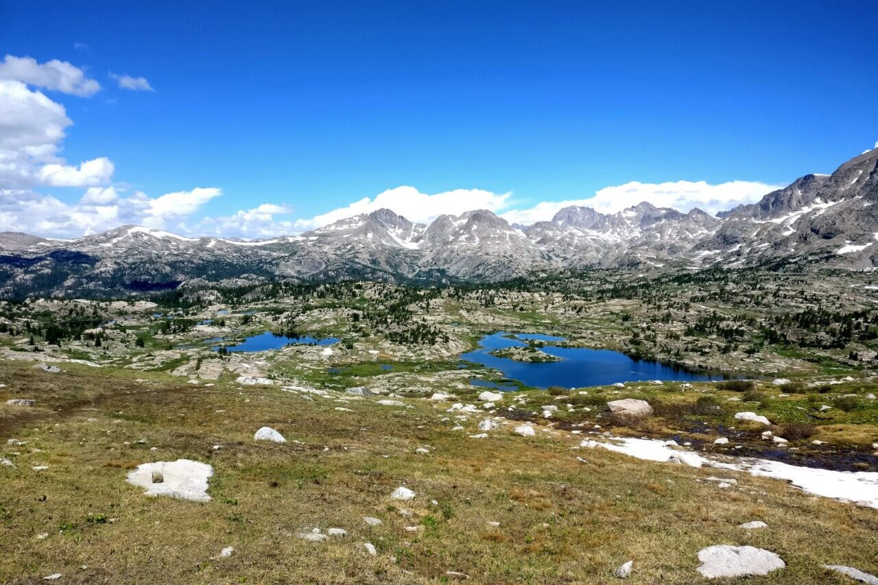 A lake sits in a rocky green and gold field in front of distant mountains.