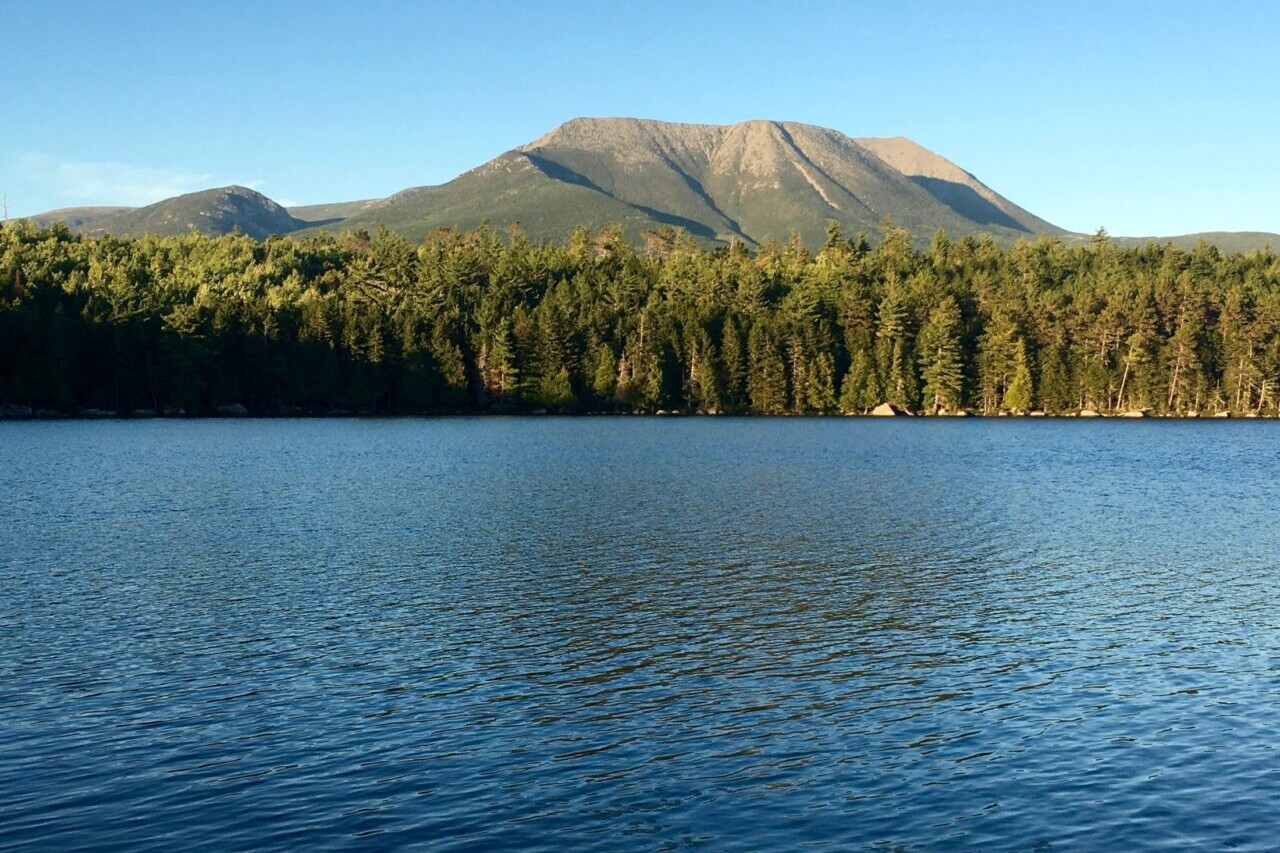 A clear lake reflects the forest and mountains surrounding it.