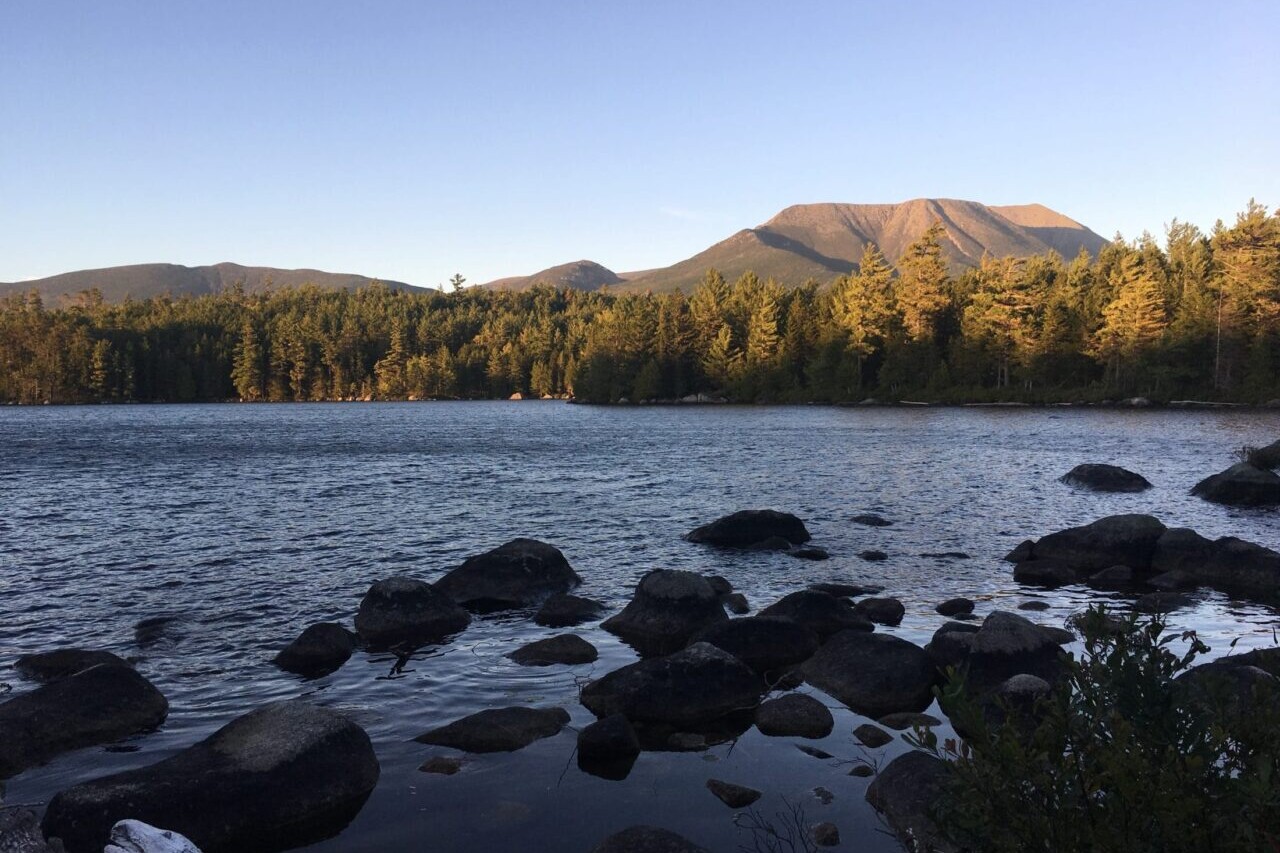 A clear lake with boulders in it reflects the forest and mountains surrounding it.
