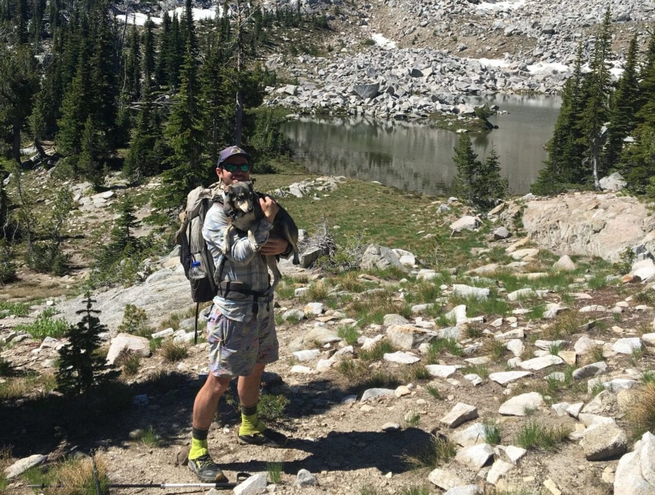 A backpacker holds a dog in front of an alpine lake.