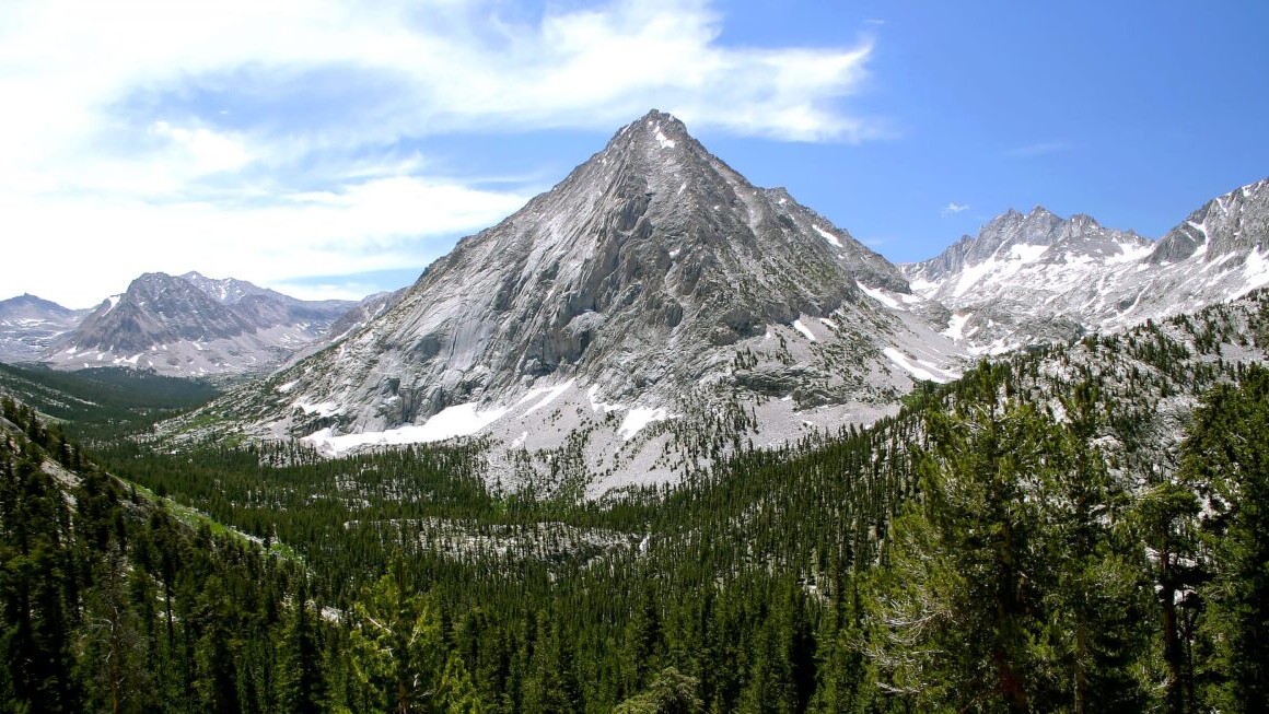 The John Muir Trail offers a commanding view of a rocky mountain peak with patchy snow surrounded by an evergreen forest.