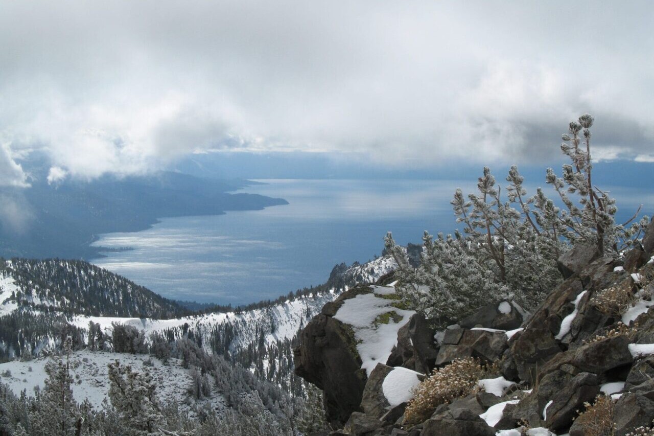 A rocky view overlooks a blue lake and meadows under a cloudy sky.