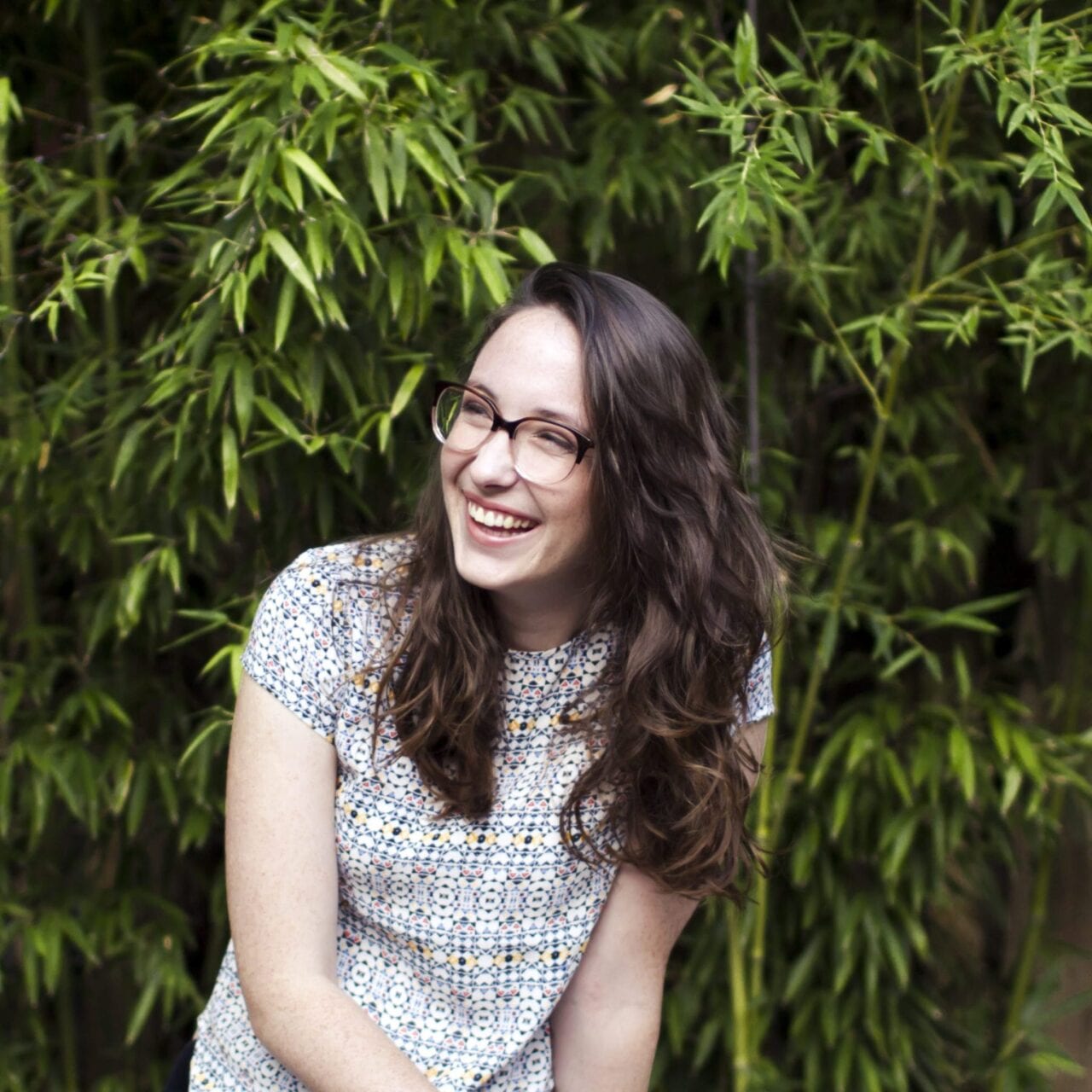 A woman wearing glasses sits in front of a bamboo grove.