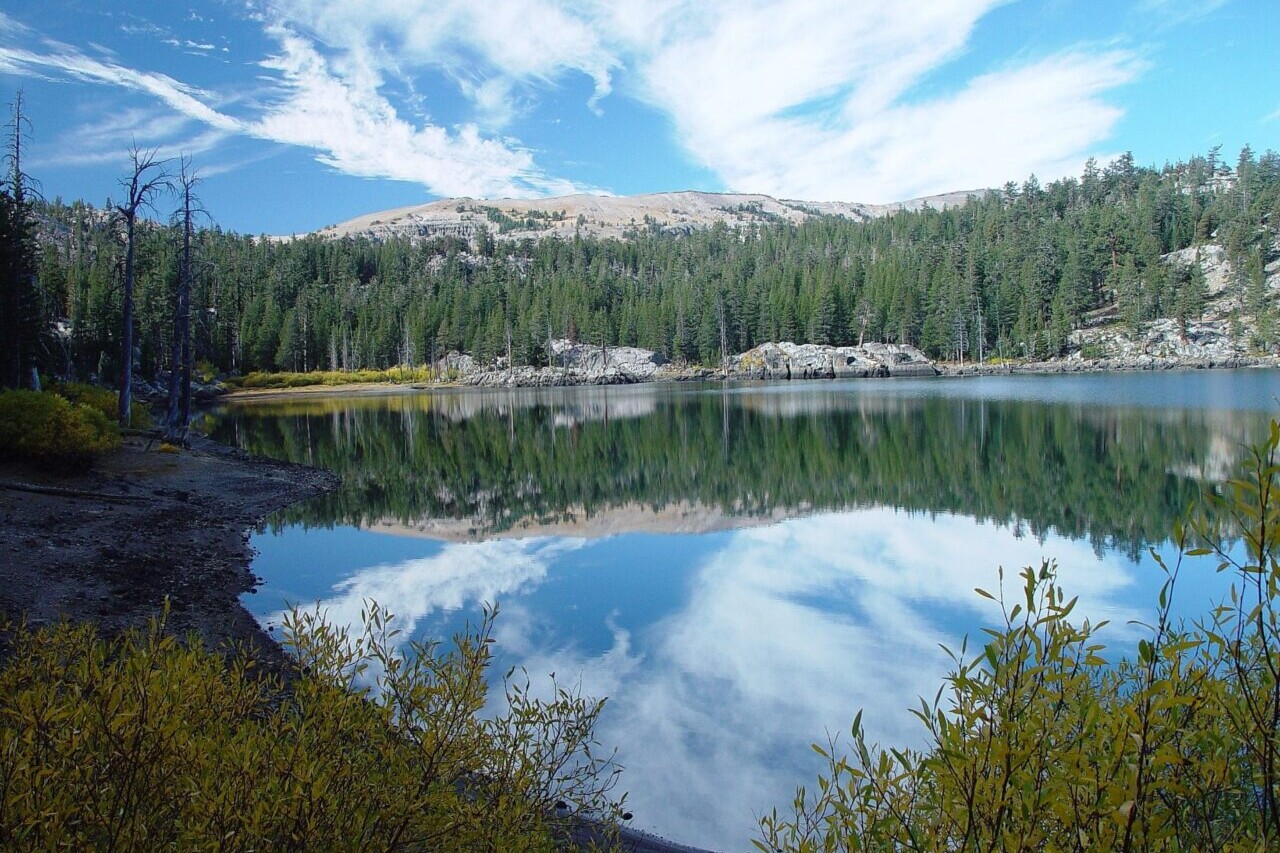 A lake reflects a nearby mountain, trees, and a blue sky.