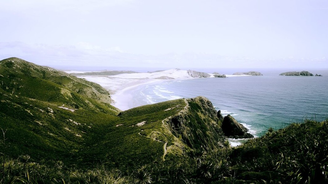 A view from Cape Reinga on the Te Araroa on the North Island of New Zealand looks over beautiful blue ocean, white sand beaches, and green hills.