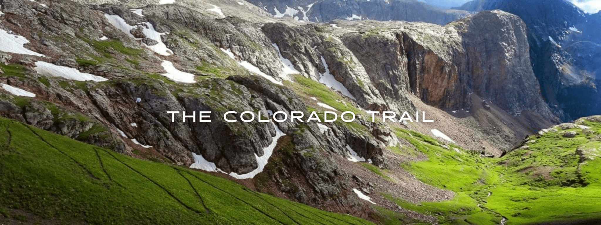 A trail winds down a steep slope to a lush green valley in between tall mountains on the Colorado Trail.