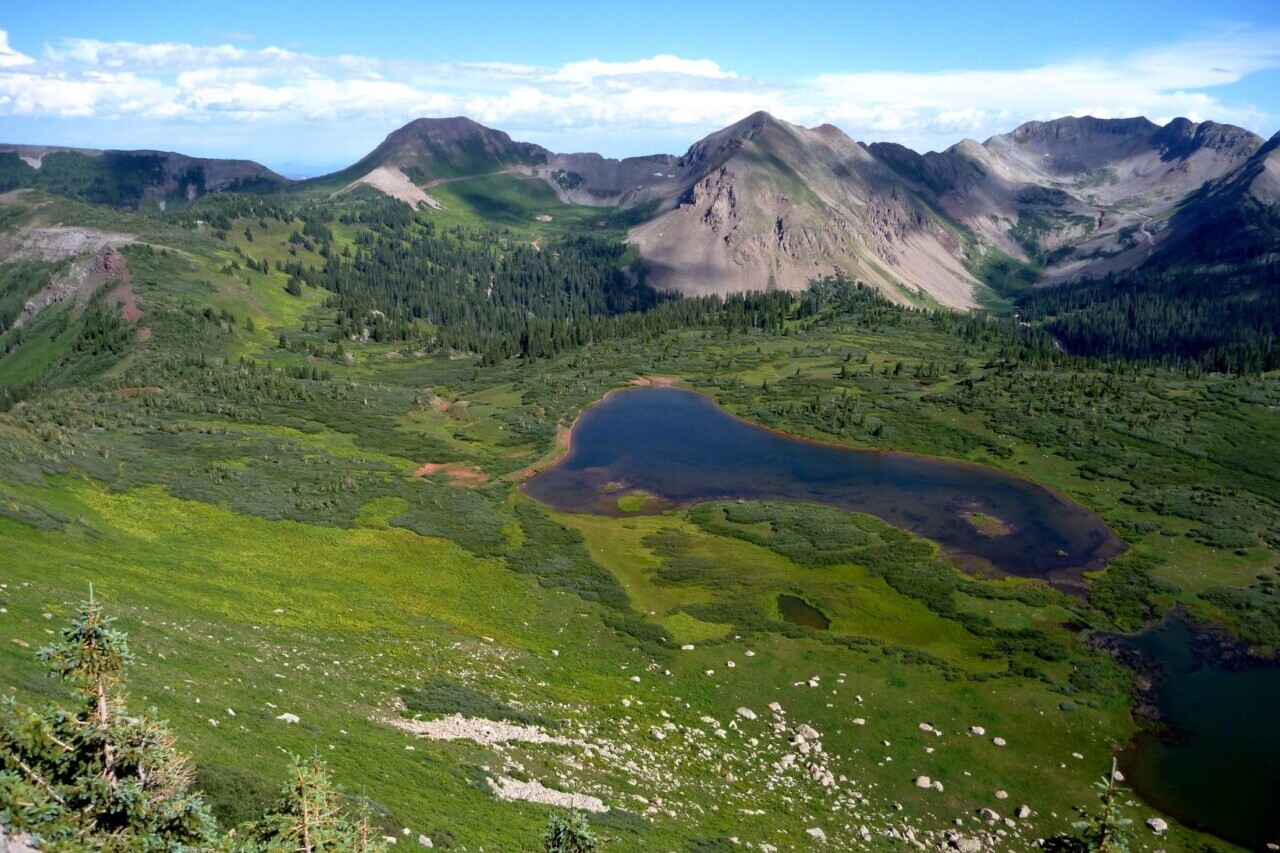 A view looks over a blue lake in a green valley surrounded by rocky mountains.