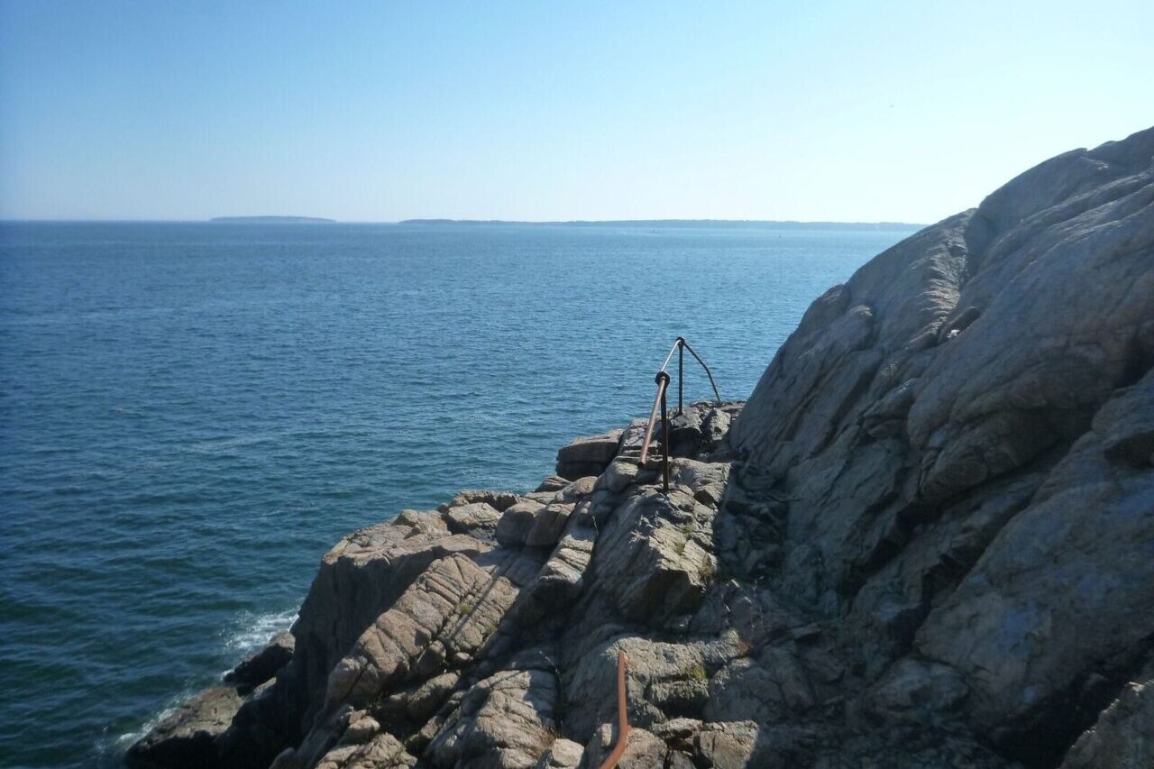 A trail winds around a rock outcropping on the edge of a large blue expanse of water.