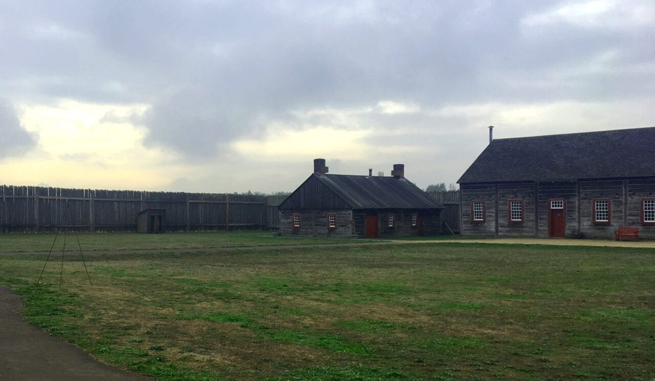 Two wooden buildings sit on a green field within a wooden wall at Fort vancouver National Historic Site.