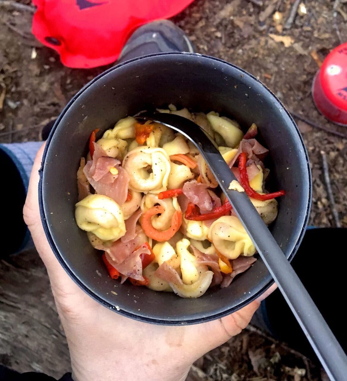 A hiker holds a pot of tortellini and vegetables with a spoon in it.