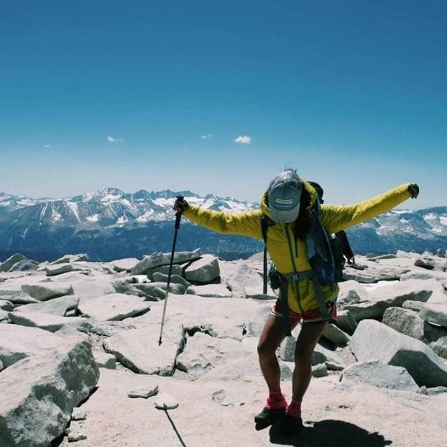 A thru hiker stands on top of a mountain with her arms up.