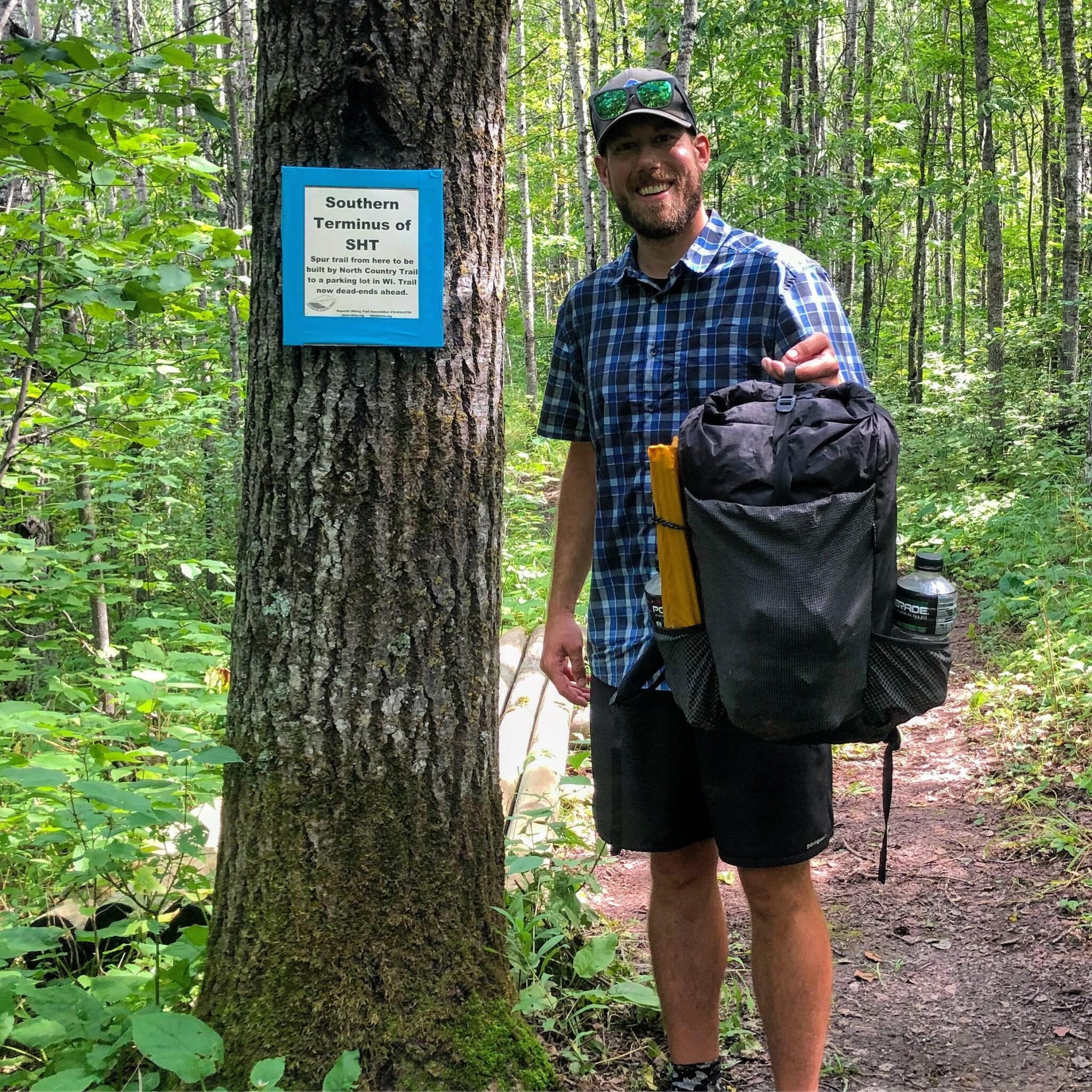 A man with a backpack standing next to a tree on a trail.