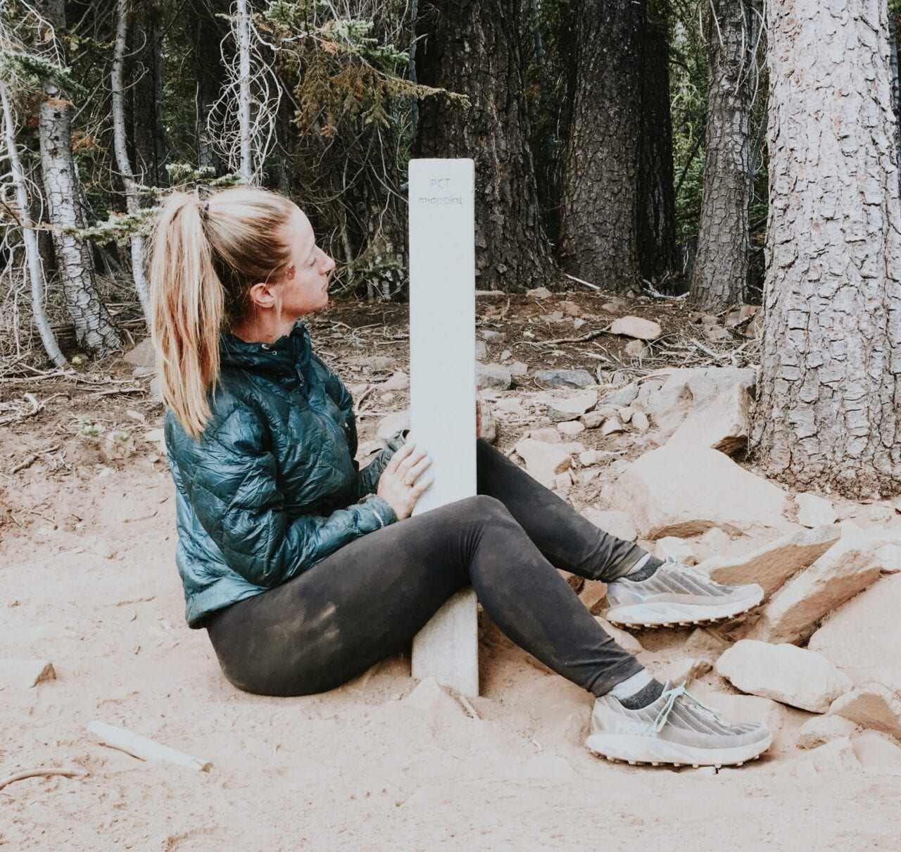 A girl sits on the ground next to a trail sign post.