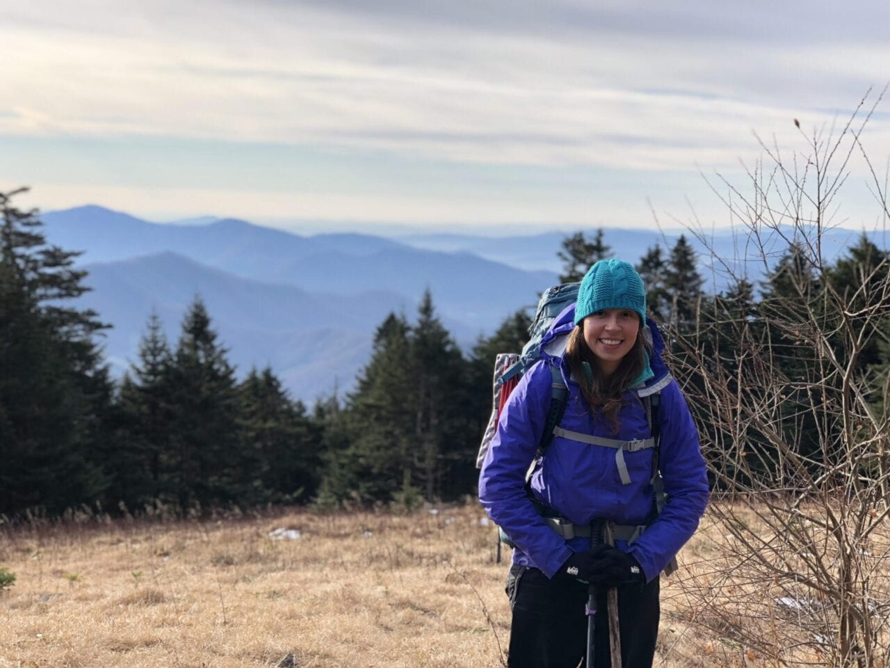 A girl hiking the Appalachian Trail stands smiling with the mountains and trees behind her.