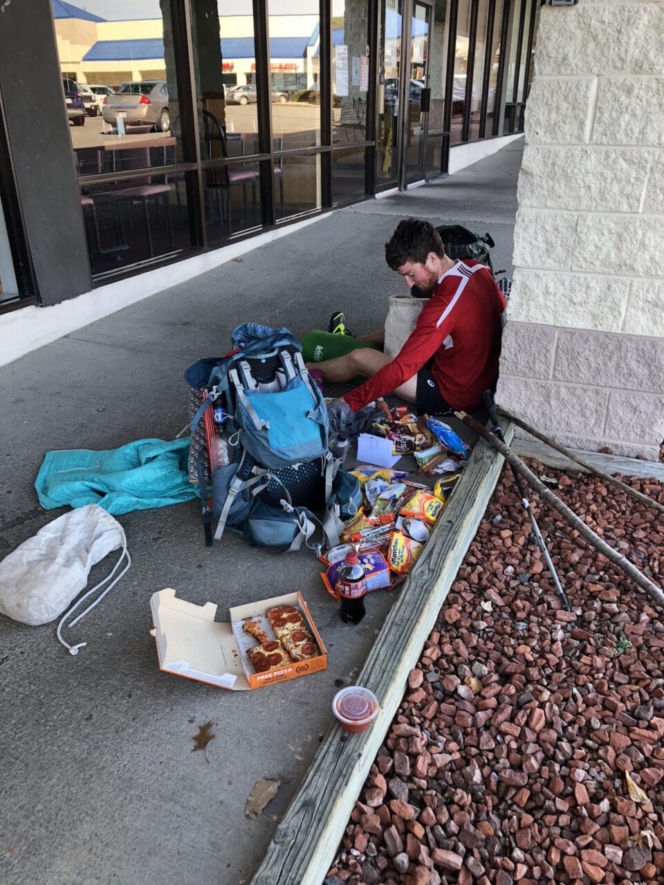 A thru-hiker sits on the ground eating with food spread out on the floor.