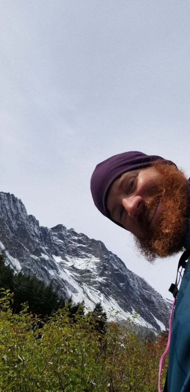 A man with a beard and a beanie smiles with a mountain behind him.