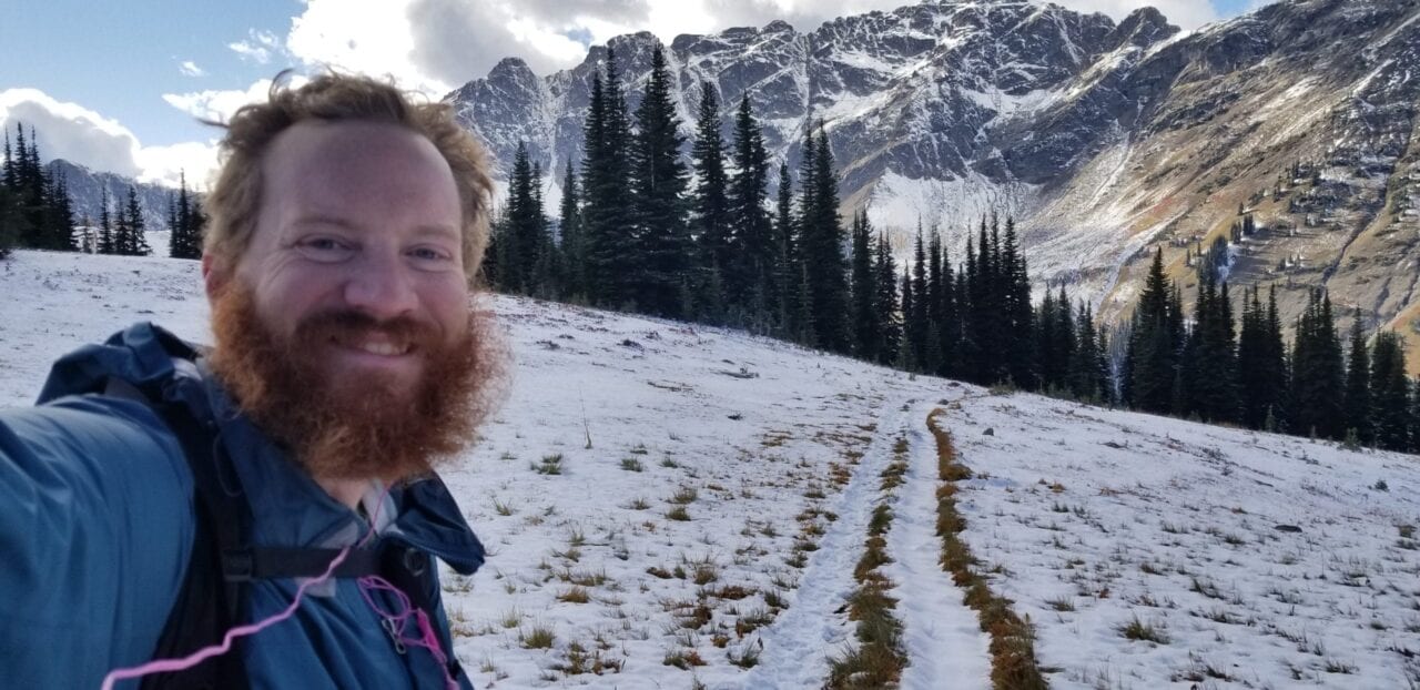 A man with a beard smiling with snow and trees behind him.