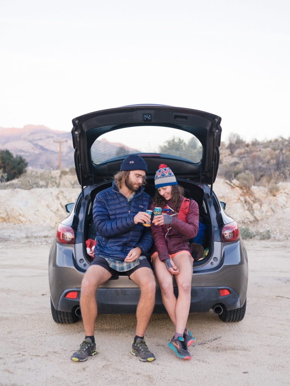 A man and woman sit in the trunk of a car cheers-ing their beers.
