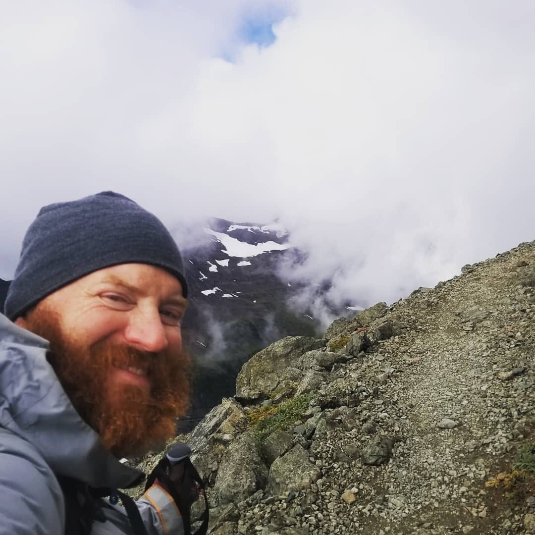 A man with a beard smiling on a mountain with clouds behind him.