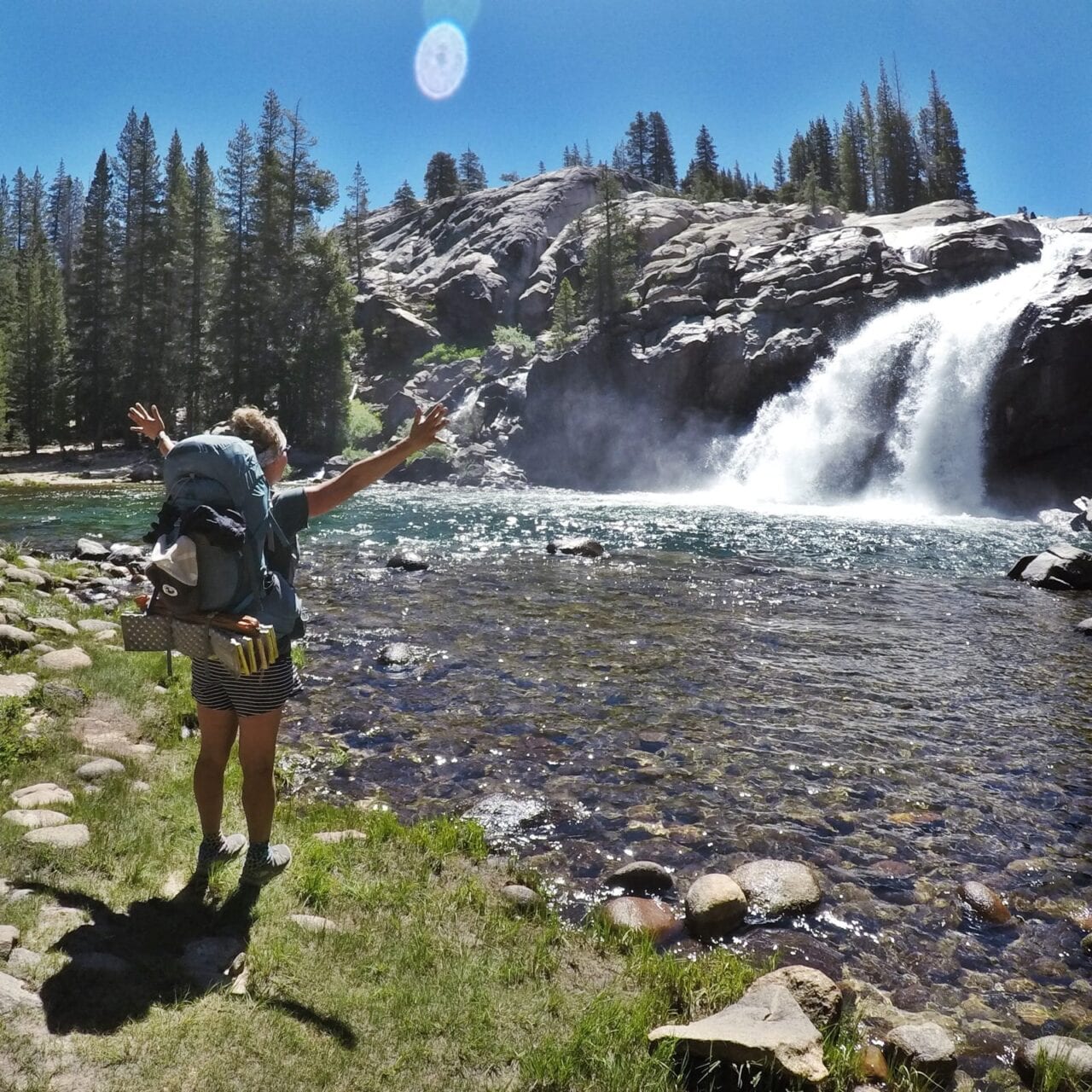 A woman hiker standing with her arms up in front of a waterfall.