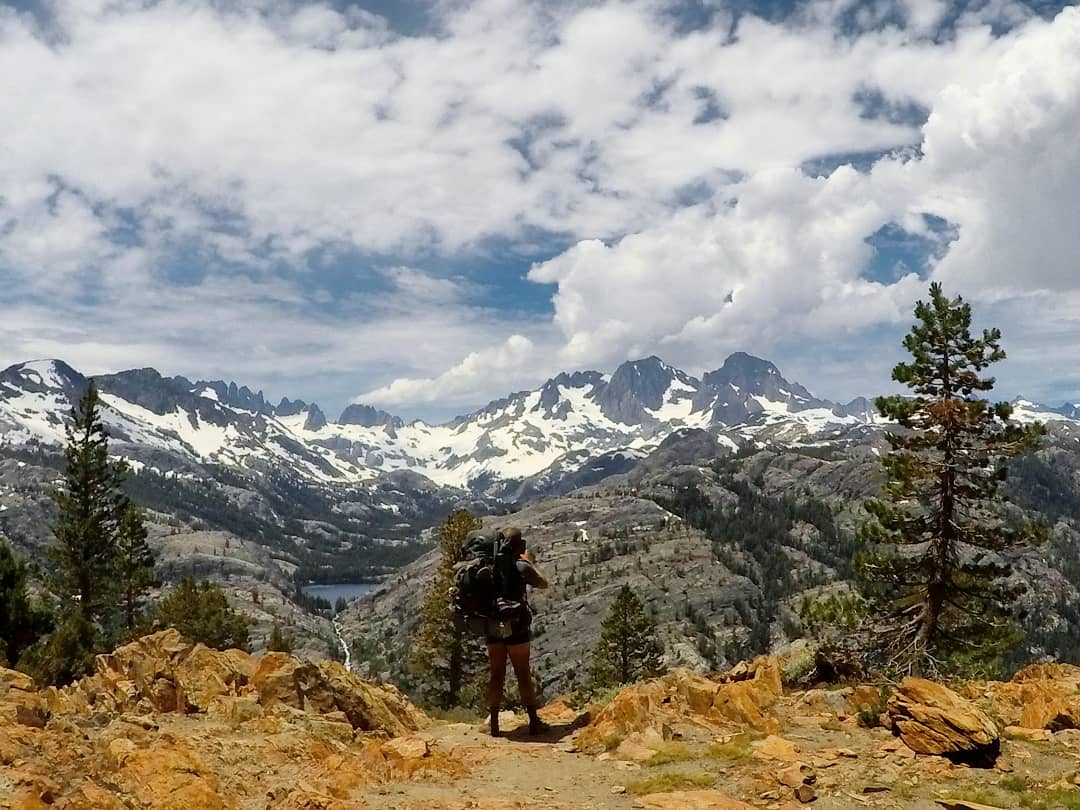 A hiker stands with a view of the mountain s behind.
