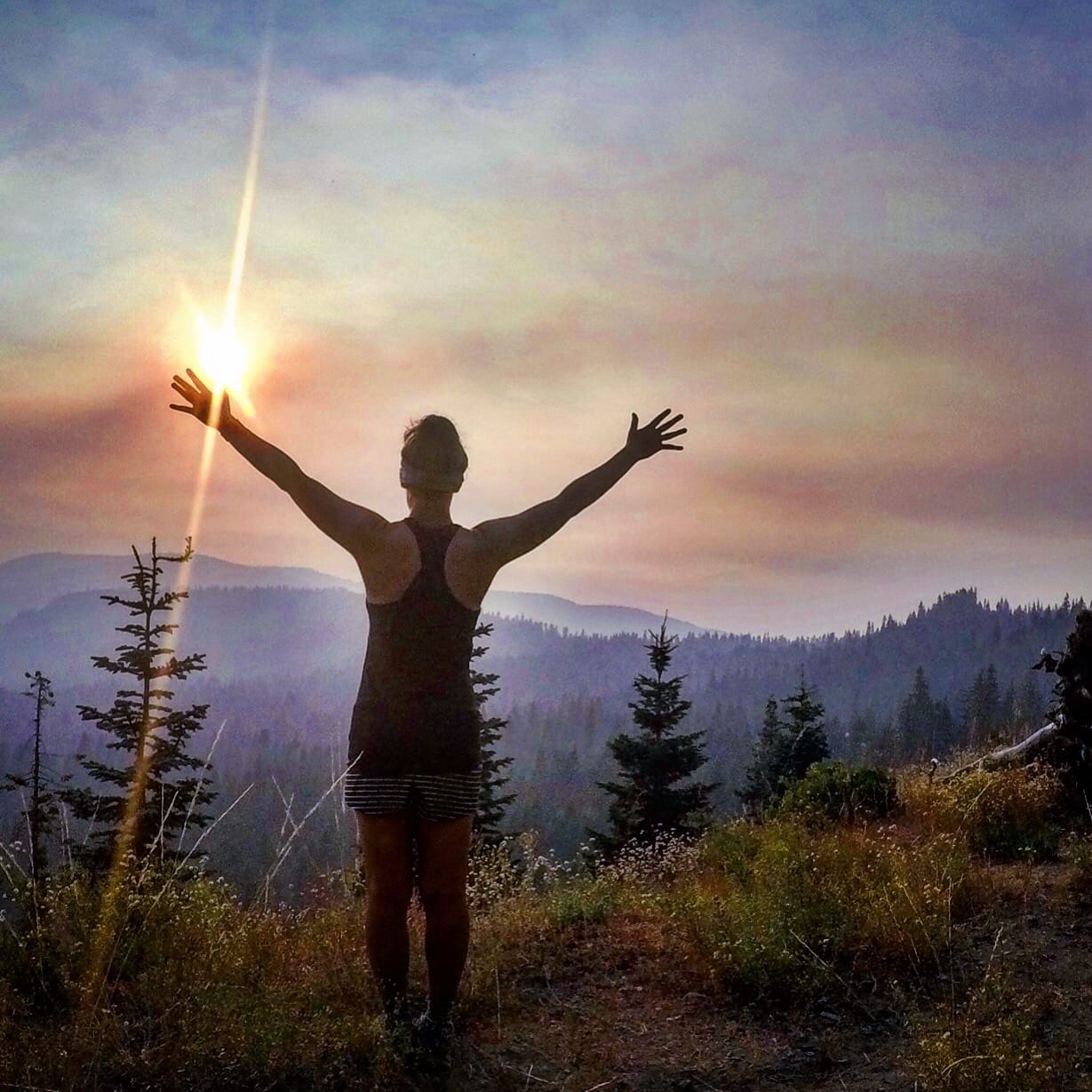 A woman standing with her arms up in front of a sunset.
