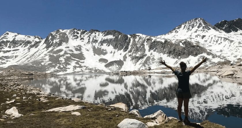 A woman stands in front of a lake by some mountains.