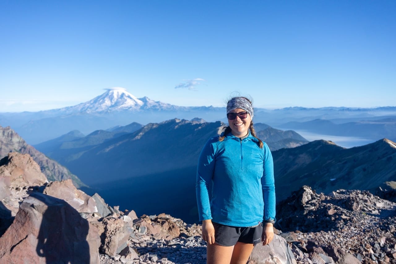 A girl stands on a mountain with a snow-capped mountain behind her.