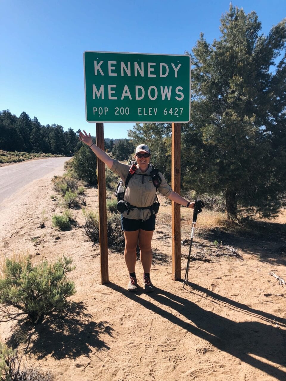 A woman stands next to a Kennedy Meadows sign with her hiking backpack and trekking poles.