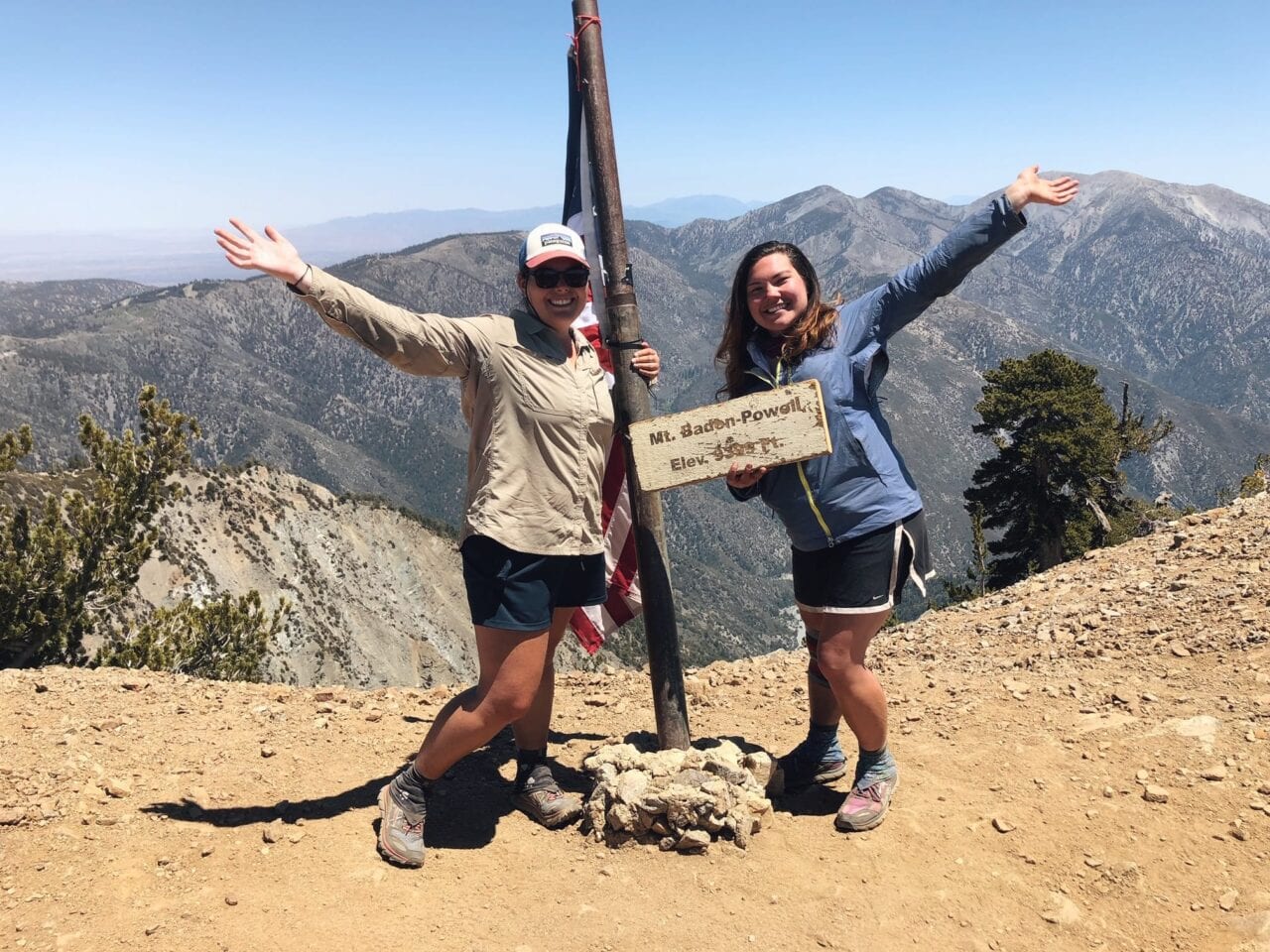 Two women standing next to a flag pole on a mountain holding a sign.