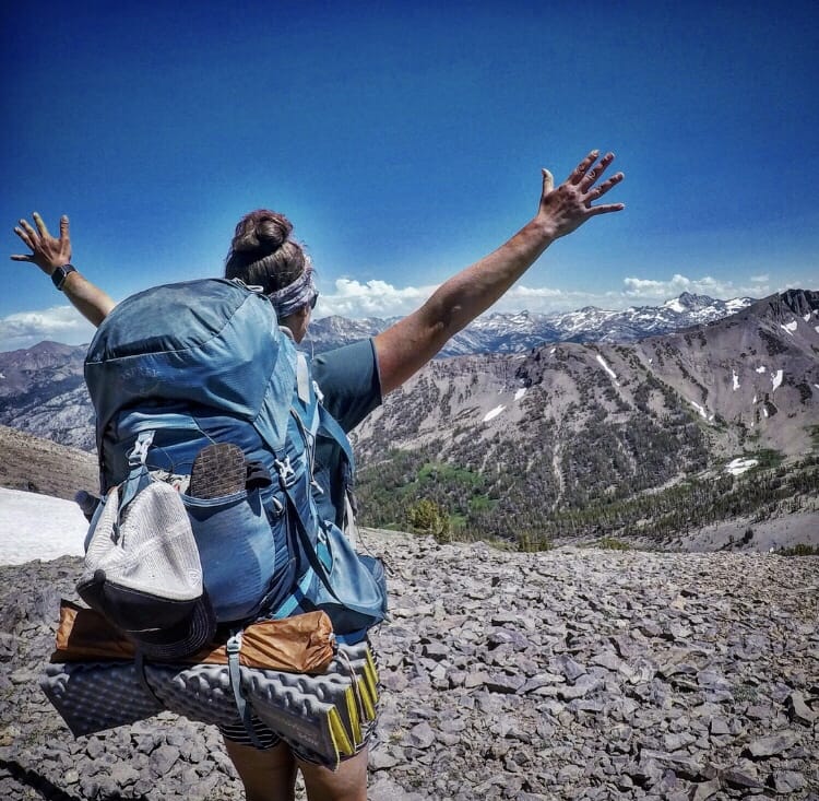 A girl hiker standing with her arms up looking out at the mountains.