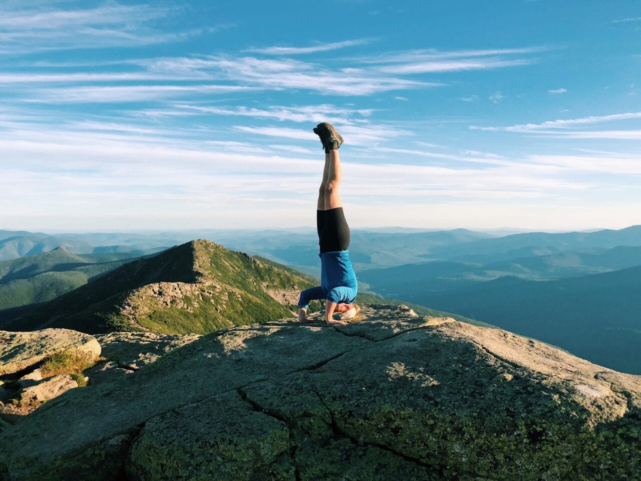 A woman doing a headstand on a hill with beautiful mountains behind her.